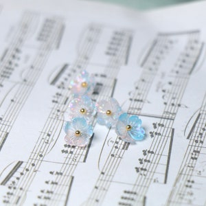 May include: Six iridescent blue and white flower-shaped resin charms with gold accents. The charms are scattered on a sheet of music.