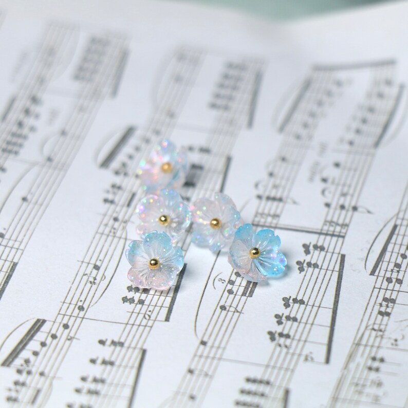May include: Six iridescent blue and white flower-shaped resin charms with gold accents. The charms are scattered on a sheet of music.
