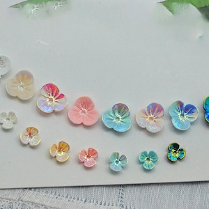 Puede incluir: Surtido de adornos iridiscentes en forma de flor en varios colores, incluyendo rosa, azul y blanco. Los adornos son perfectos para agregar un toque de brillo a las manualidades y proyectos.