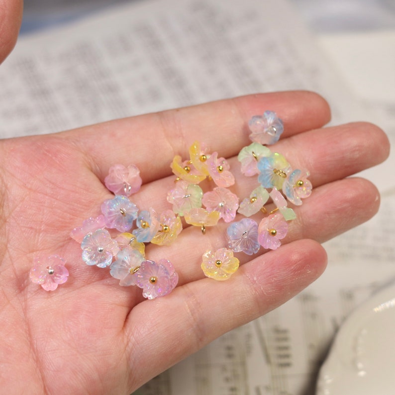 May include: A collection of pastel-colored flower-shaped resin charms with gold-colored wire loops. The charms are scattered on a hand.