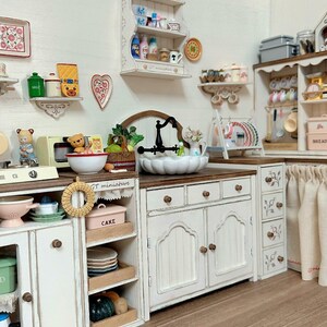 Pu&ograve; includere: Una scena di cucina in miniatura con mobili, mensole e un lavandino bianchi. La scena comprende vari oggetti da cucina in miniatura come piatti, tazze e generi alimentari. Il testo "Display only. Props not included" &egrave; visibile in alto.