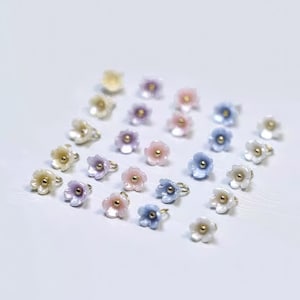 Könnte beinhalten: Eine Sammlung kleiner, blütenförmiger Ringe in verschiedenen Pastellfarben, darunter Weiß, Rosa, Lila und Blau. Jeder Ring hat eine goldfarbene Mitte und ein Band. Die Ringe sind auf einer weißen Oberfläche angeordnet.