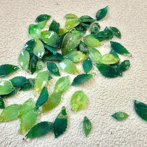 May include: A collection of green glass leaves in various shades, from deep emerald to light lime, scattered on a textured, cream-coloured surface. The leaves have a glossy finish and some appear to have water droplets.