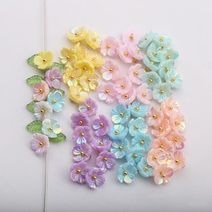 May include: Assortment of pastel colored iridescent flower shaped resin embellishments with gold accents. The flowers are arranged in rows and are perfect for crafting and jewelry making.