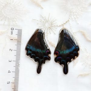 Puede incluir: Dos colgantes iridiscentes con forma de alas de mariposa, con una base oscura y reflejos azules, verdes y morados. Los colgantes están junto a una regla de centímetros, sobre un fondo blanco texturizado.