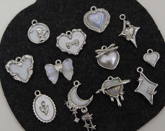 May include: A collection of 12 silver charms with white enamel and pearl accents. The charms feature various designs, including hearts, stars, moons, butterflies, and angels.