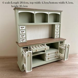 Op de afbeelding: Een miniatuur saliegroene kast met meerdere lades en planken. De kast heeft een houten aanrechtblad en witte accenten. De afmetingen zijn: lengte 28,6 cm, bovenbreedte 4,3 cm, onderbreedte 8,5 cm en hoogte 30,5 cm. De kast heeft een klassiek ontwerp.