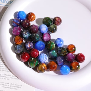 May include: A collection of colorful, iridescent glass beads. The beads are round and have a variety of colors, including blue, green, red, and gold. The beads are scattered on a white surface.