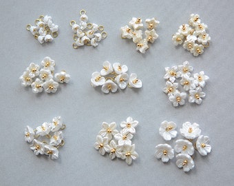 Puede incluir: Surtido de abalorios de flores de perlas blancas con detalles dorados. Los abalorios están dispuestos en grupos de 5 a 10 sobre una superficie gris claro.