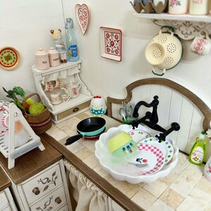 Pu&ograve; includere: Scena di cucina in miniatura con un lavandino bianco, piatti e un rubinetto nero. Sono visibili anche una piccola padella, un cestino di verdure e oggetti decorativi come un servizio da t&egrave; e decorazioni murali.