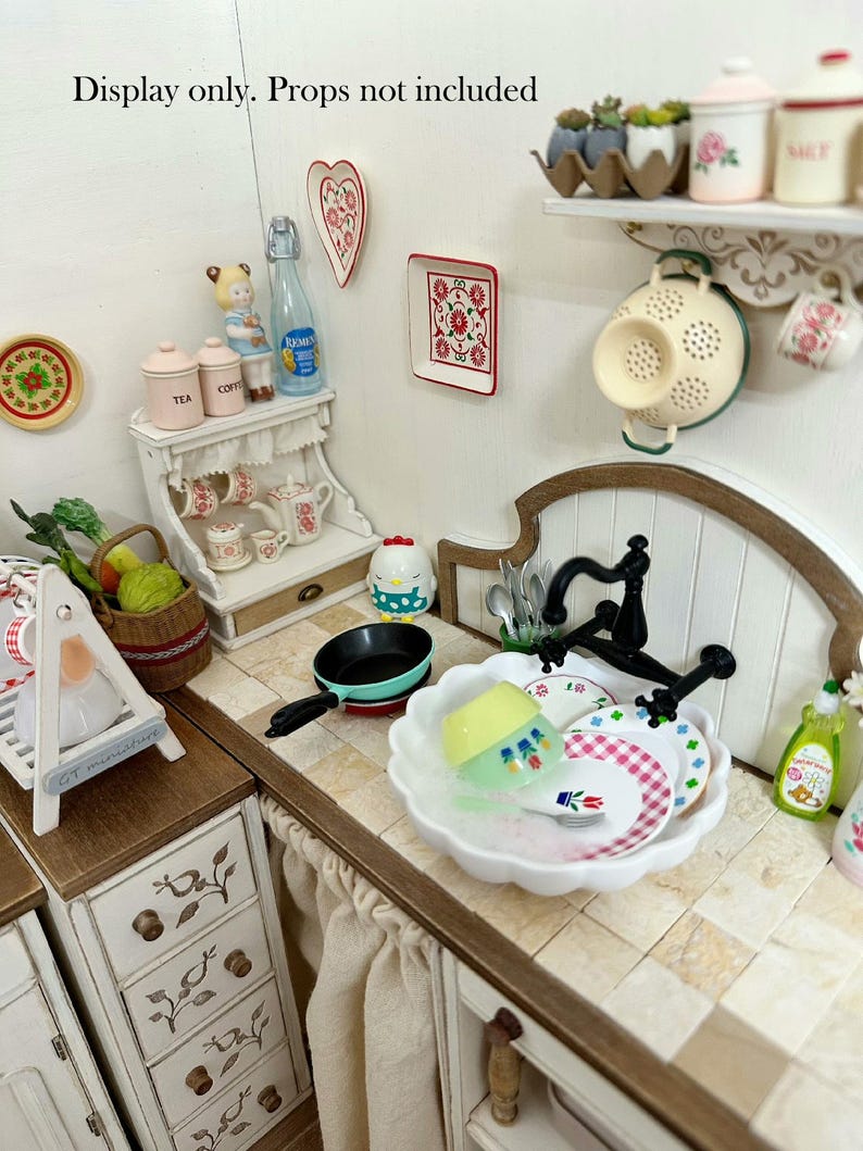Pu&ograve; includere: Scena di cucina in miniatura con un lavandino bianco, piatti e un rubinetto nero. Sono visibili anche una piccola padella, un cestino di verdure e oggetti decorativi come un servizio da t&egrave; e decorazioni murali.