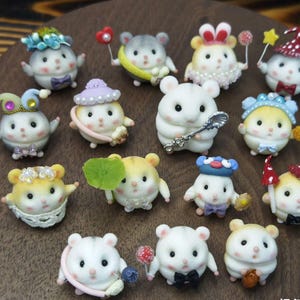 May include: A selection of handcrafted hamster figurines. The hamsters are white, cream, or yellow, and decorated with miniature hats, bows, and accessories. Some hold small props such as spoons or wands, creating a detailed and charming display.