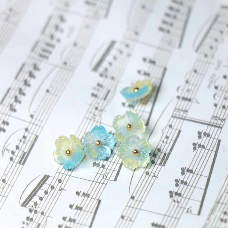 May include: Five pastel blue and yellow flower-shaped resin embellishments with gold accents on a sheet of music.
