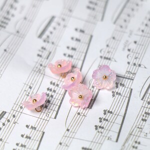 May include: Five pink flower-shaped buttons with gold accents and a loop on the back. The buttons are arranged on a sheet of music.