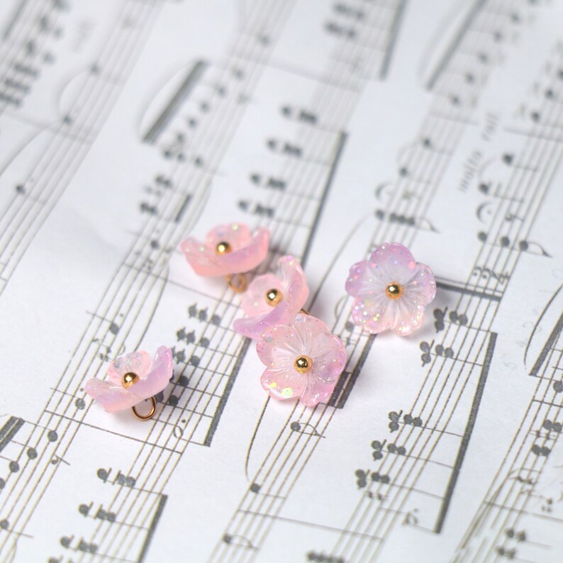 May include: Five pink flower-shaped buttons with gold accents and a loop on the back. The buttons are arranged on a sheet of music.