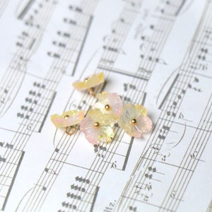 May include: Five iridescent pastel pink and yellow flower-shaped buttons with gold centers. The buttons are arranged on a sheet of music.