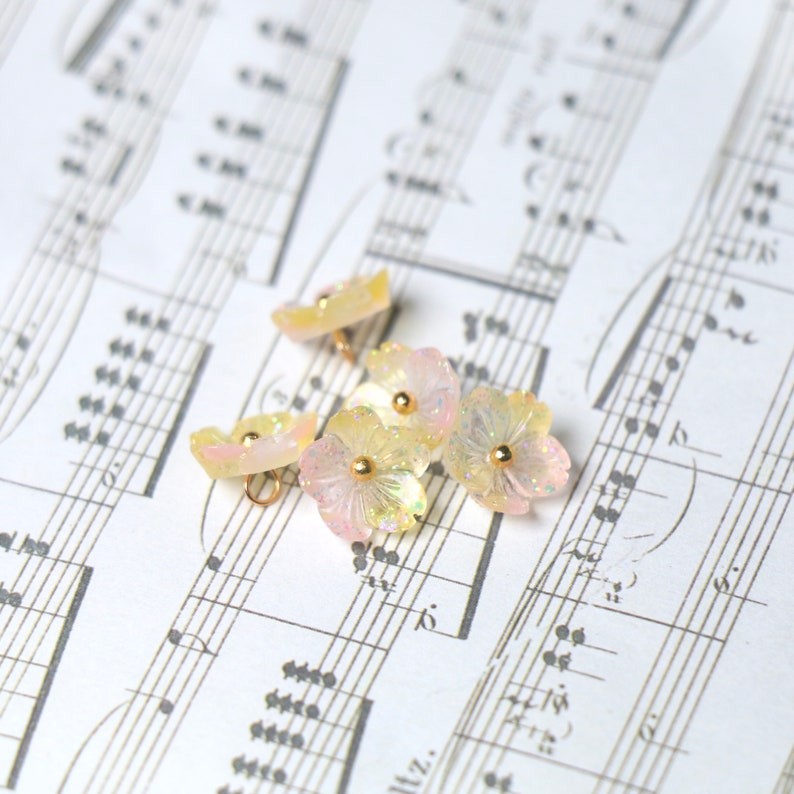 May include: Five iridescent pastel pink and yellow flower-shaped buttons with gold centers. The buttons are arranged on a sheet of music.