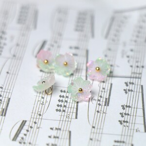 May include: Five iridescent pastel pink and green flower-shaped buttons with gold accents. The buttons are scattered on a sheet of music.
