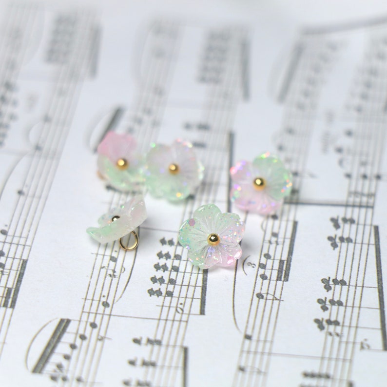 May include: Five iridescent pastel pink and green flower-shaped buttons with gold accents. The buttons are scattered on a sheet of music.