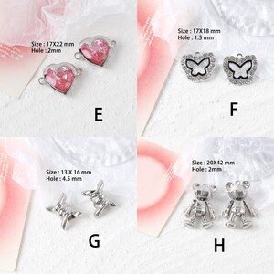 May include: Assortment of silver-toned metal charms with various designs.  The charms include a heart with pink glitter, a butterfly with rhinestones, a barbed wire design, and a robot-like figure.  The charms are displayed on a white background.
