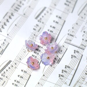 May include: Six pastel purple and pink flower charms with gold accents. The charms are scattered on a sheet of music.