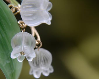 May include: Close-up of a delicate glass lily of the valley flower charm with a gold chain. The charm is white and translucent, with a detailed design.