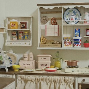 Pu&ograve; includere: Una scena di cucina in miniatura con mensole bianche piene di generi alimentari, tra cui barattoli, lattine e scatole. Un piccolo fornello con pentole di rame &egrave; appoggiato sul bancone. La scena comprende una scatola per il pane, piatti e oggetti decorativi.