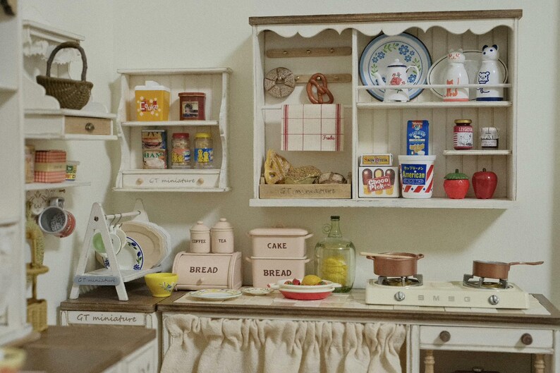 Pu&ograve; includere: Una scena di cucina in miniatura con mensole bianche piene di generi alimentari, tra cui barattoli, lattine e scatole. Un piccolo fornello con pentole di rame &egrave; appoggiato sul bancone. La scena comprende una scatola per il pane, piatti e oggetti decorativi.