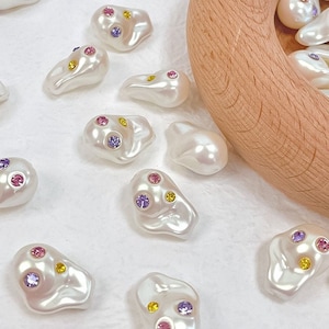 Puede incluir: Una colección de cuentas blancas con forma de perla con piedras de imitación de colores. Las cuentas están esparcidas sobre una superficie blanca y en un cuenco de madera.