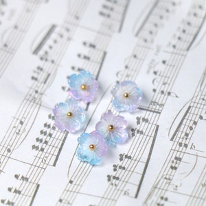 May include: Five pastel blue and purple flower-shaped resin embellishments with gold centers. The embellishments are scattered on a sheet of music.