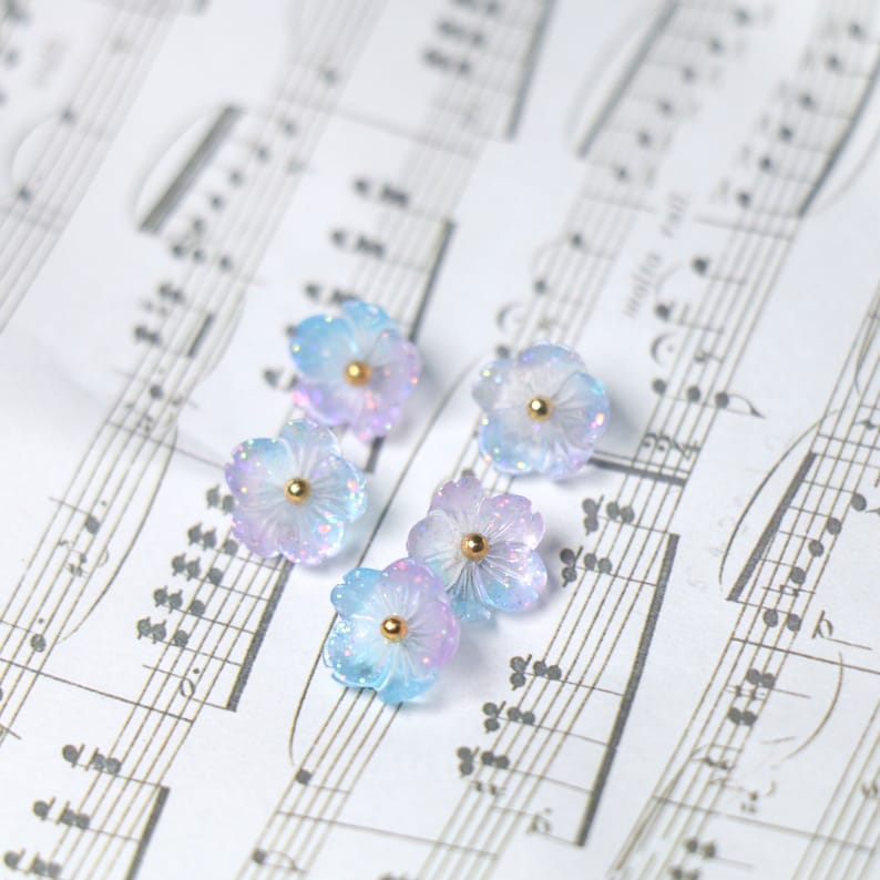 May include: Five pastel blue and purple flower-shaped resin embellishments with gold centers. The embellishments are scattered on a sheet of music.