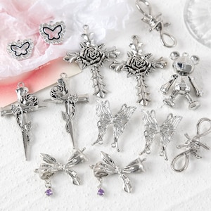 May include: A collection of silver-toned metal charms featuring various designs, including crosses with roses, butterflies, bows, a bear, and a barbed wire loop.