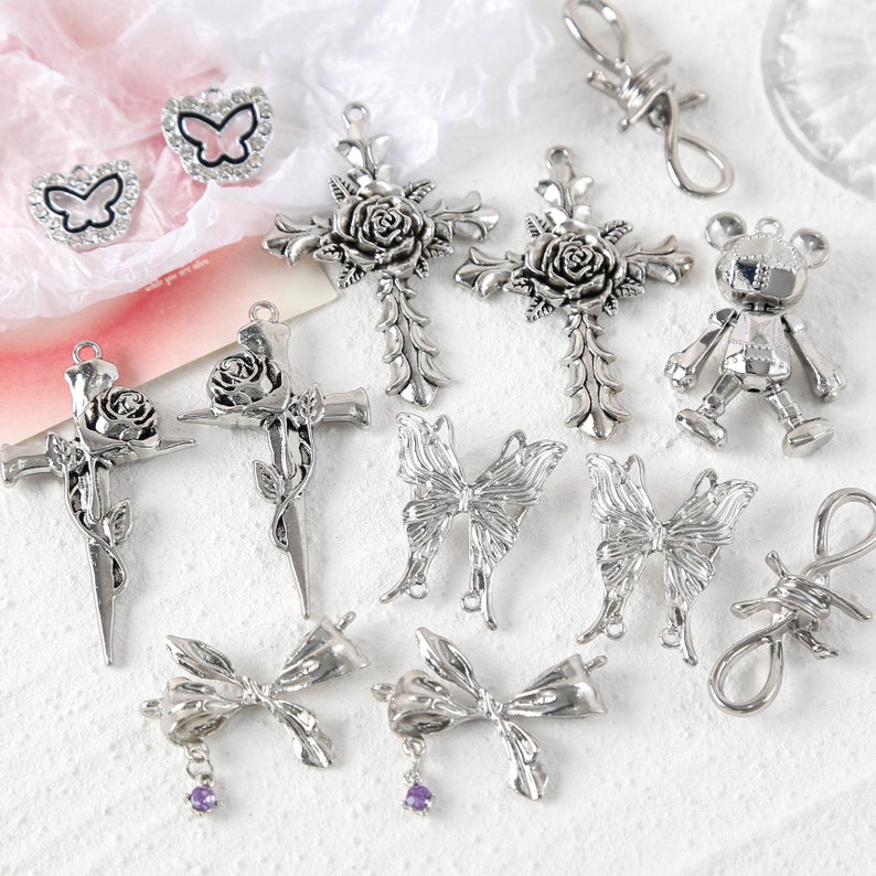 May include: A collection of silver-toned metal charms featuring various designs, including crosses with roses, butterflies, bows, a bear, and a barbed wire loop.