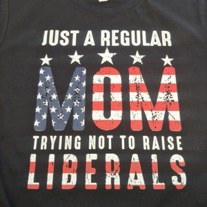 May include: Black t-shirt with white text that reads "Just a regular mom trying not to raise liberals". The word "mom" is in large letters and is styled like the American flag.