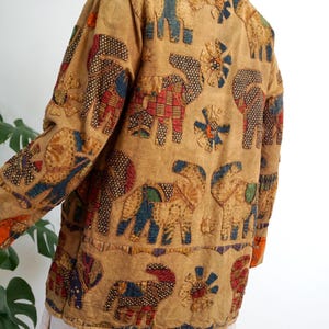 May include: A tan jacket featuring a repeating pattern of appliqued elephants and circular floral motifs. The elephants are constructed from various colorful fabric patches in shades of red, blue, and green with diverse geometric patterns.