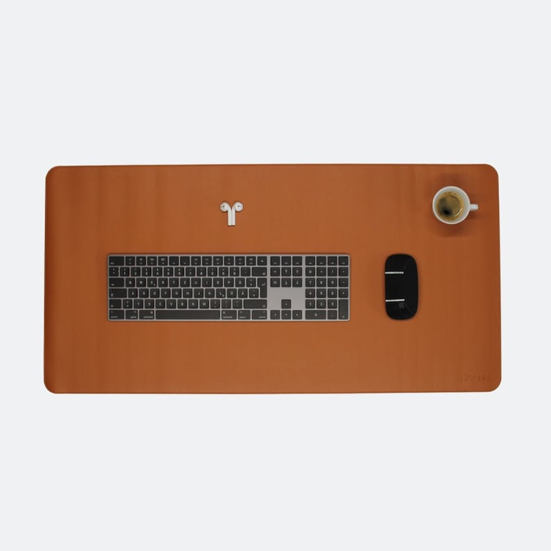 Ganze Matte ist zusehen in der Farbe cognac. Auf der Matte steht oben rechts ein Espresso. In der Mitte ist ein Magic Keyboard und mitte/rechts liegt eine Maustaste. Unten rechts steht das Logo NESK. Oben mittig liegen Airpods.