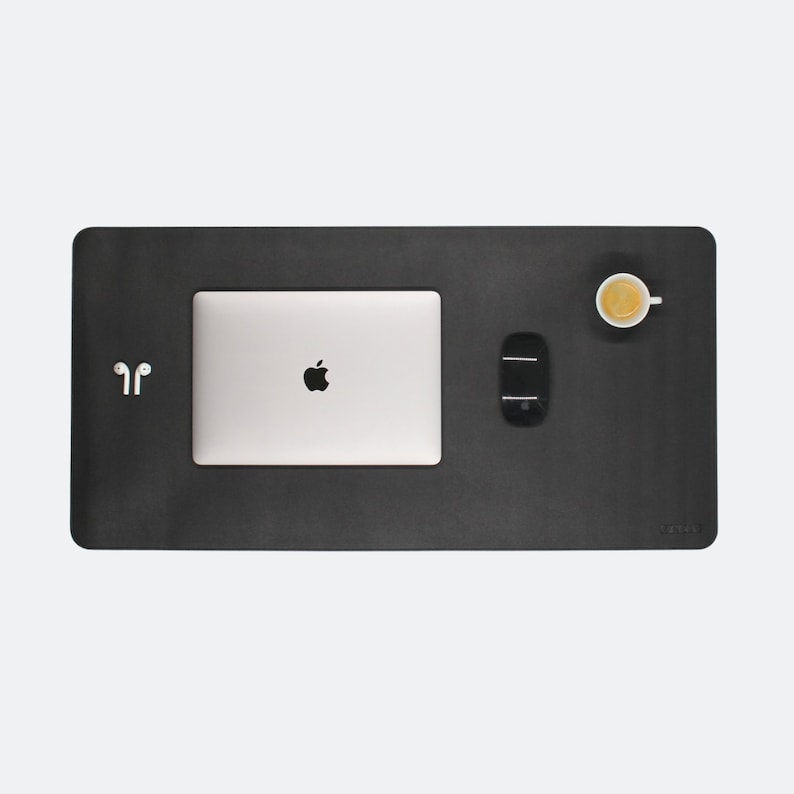 Ganze Matte ist zusehen in der Farbe schwarz zu. Auf der Matte steht oben rechts ein Espresso. In der Mitte ist ein  Mac Book und mitte/rechts liegt eine Maustaste. Unten rechts steht das Logo NESK. Mitte links liegen Airpods.