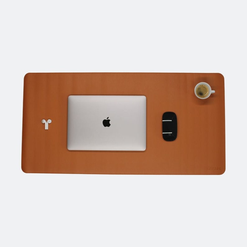 Ganze Matte ist zusehen in der Farbe cognac. Auf der Matte steht oben rechts ein Espresso. In der Mitte ist ein  Mac Book und mitte/rechts liegt eine Maustaste. Unten rechts steht das Logo NESK. Mitte links liegen Airpods.