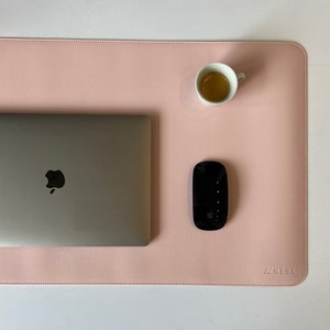 Halbe Matte ist zusehen in der Farbe rosé. Auf der Matte steht oben rechts ein Espresso. In der Mitte ist ein halber Mac Book und mitte/rechts liegt eine Maustaste. Unten rechts steht das Logo NESK