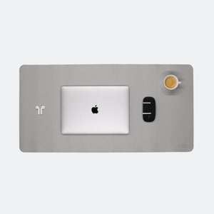 Ganze Matte ist zusehen in der Farbe steingrau. Auf der Matte steht oben rechts ein Espresso. In der Mitte ist ein  Mac Book und mitte/rechts liegt eine Maustaste. Unten rechts steht das Logo NESK. Mitte links liegen Airpods.