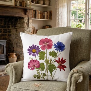 May include: A square white throw pillow featuring a botanical illustration of various flowers including a purple daisy, a pink flower, a blue cornflower, and a red flower with green leaves and stems centered on the front of the fabric cover.