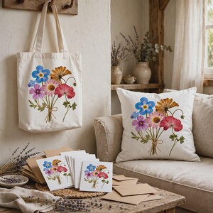 May include: A white canvas tote bag and a square throw pillow, both featuring a printed bouquet of colorful wildflowers including blue, pink, and orange blossoms. A stack of matching greeting cards with the same floral design sits on a table.