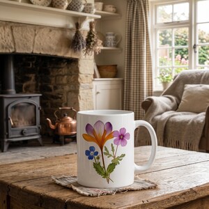May include: A white ceramic mug featuring a botanical illustration of a large orange and purple crocus, a smaller pink flower, and a tiny blue blossom with green leaves and stems, centered on the front of the mug with a curved handle.