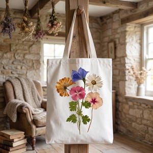 May include: A white canvas tote bag featuring a central botanical illustration of a bouquet of flowers. The design includes a blue bell-shaped flower, a white daisy, a golden-orange flower, a pink blossom, and a peach-colored flower with green leaves.