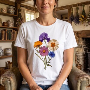 May include: A white short-sleeved t-shirt featuring a central graphic print of a colorful wildflower bouquet. The floral arrangement includes a purple iris, an orange marigold, a pink cosmos, a white daisy, and a blue cornflower with green stems.