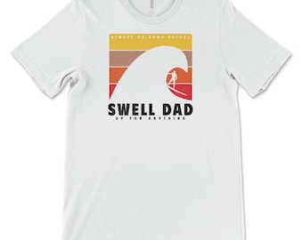 Può includere: Maglietta bianca con una grafica retrò di un surfista che cavalca un'onda. La grafica dice "Swell Dad Up For Anything" e "Always On Dawn Patrol".