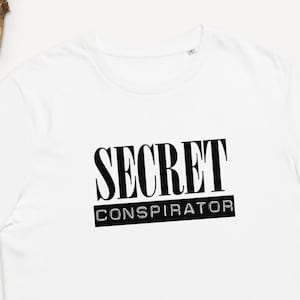Puede incluir: Camiseta blanca con texto negro que dice "SECRET CONSPIRATOR".