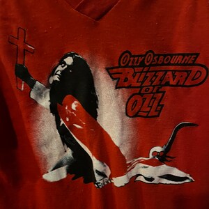 May include: Red t-shirt with a black and white image of a person holding a cross. The text "Ozzy Osbourne Blizzard of Ozz" is printed in red on the shirt.