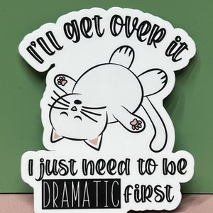 May include: White sticker with a black outline featuring a cartoon white cat lying on its back with its paws in the air. The text reads "I'll get over it I just need to be DRAMATIC first."