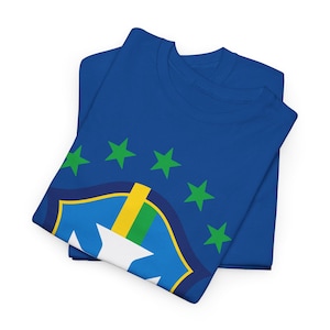Puede incluir: Dos camisetas azules dobladas con un diseño de la bandera brasileña. El diseño presenta un escudo con una estrella blanca, detalles en azul, amarillo y verde, y estrellas verdes. Las camisetas están hechas de un material suave.